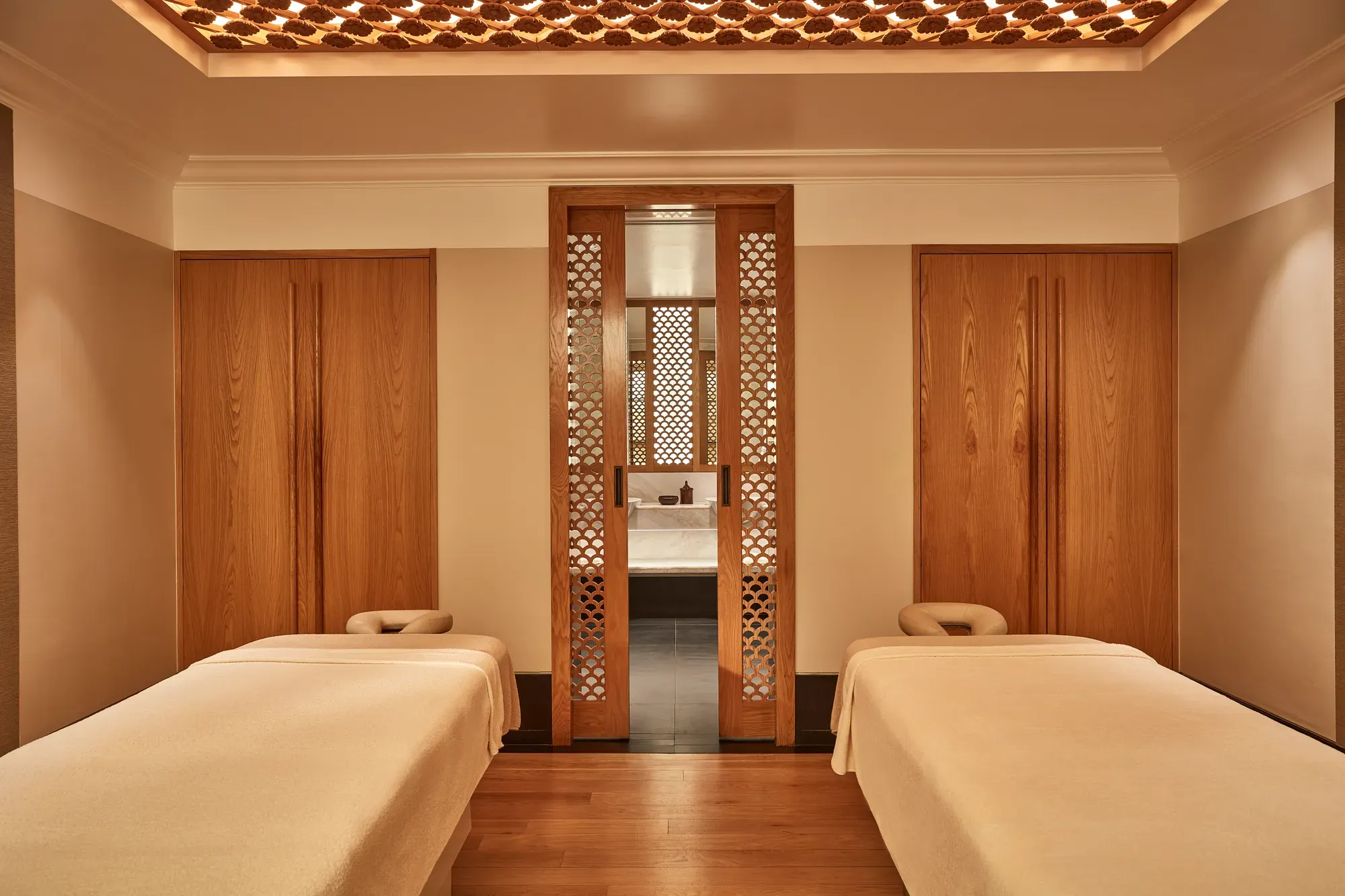 Calm spa treatment room with two massage tables, wooden lattice doors, and warm soft lighting.