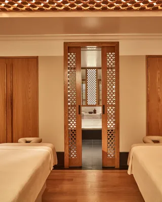 Calm spa treatment room with two massage tables, wooden lattice doors, and warm soft lighting.