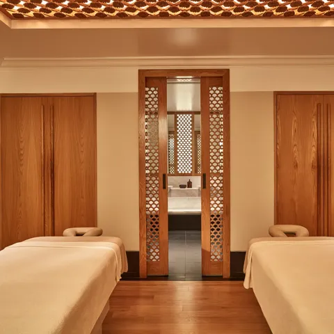 Calm spa treatment room with two massage tables, wooden lattice doors, and warm soft lighting.