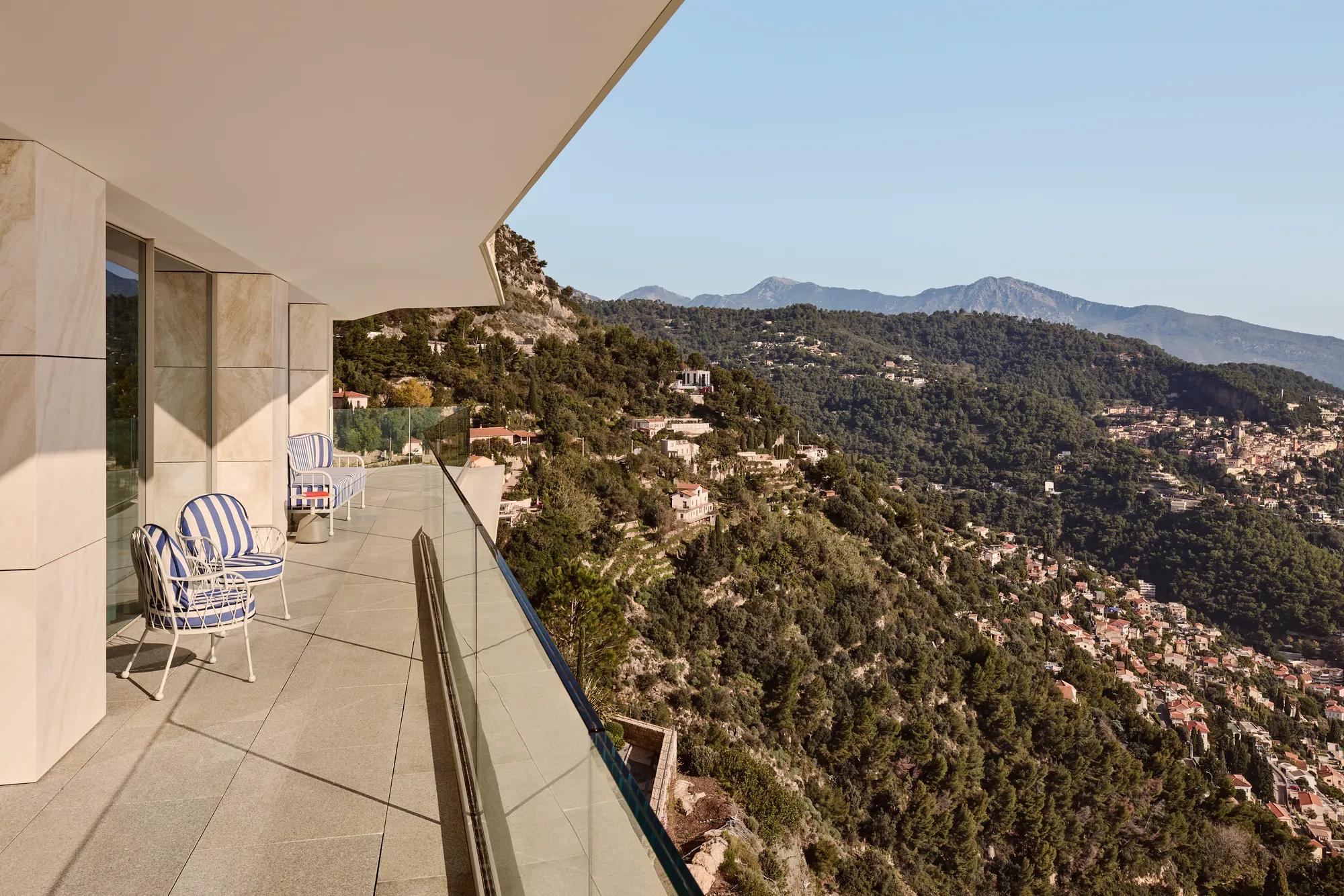 Balcon avec fauteuils rayés et garde-corps en verre, offrant une vue panoramique sur les collines boisées et un village perché sous un ciel dégagé.