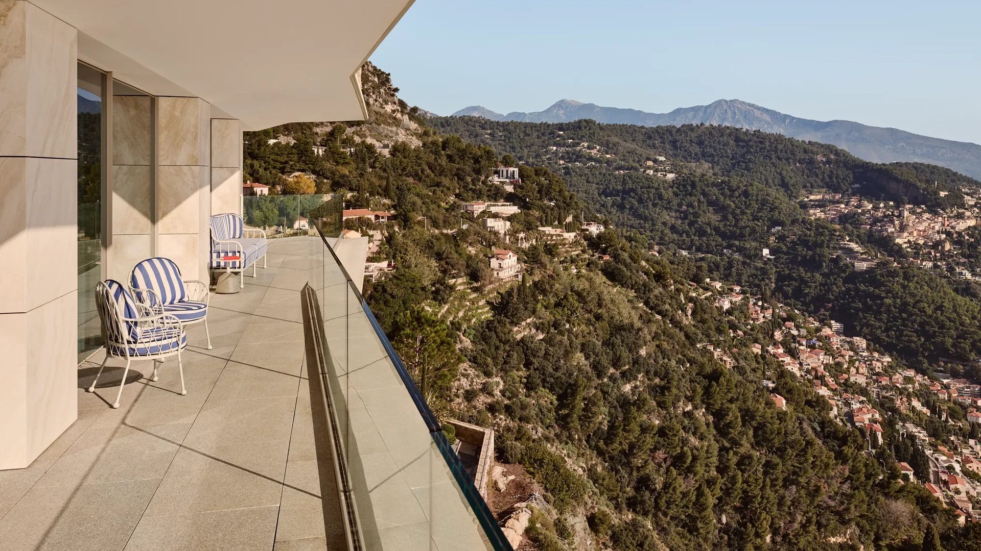 Balcon avec fauteuils rayés et garde-corps en verre, offrant une vue panoramique sur les collines boisées et un village perché sous un ciel dégagé.