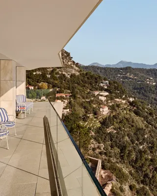 Balcon avec fauteuils rayés et garde-corps en verre, offrant une vue panoramique sur les collines boisées et un village perché sous un ciel dégagé.