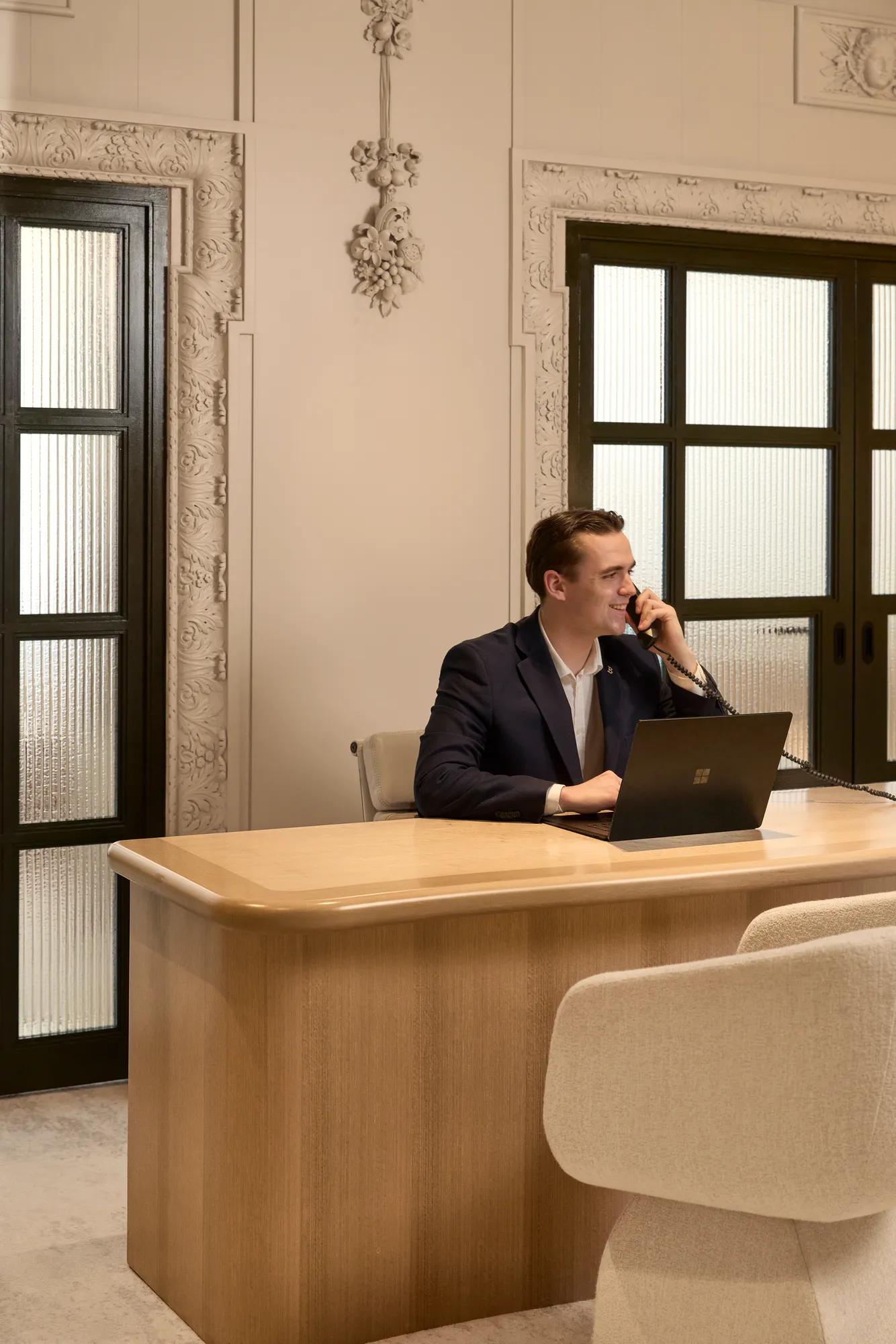 Man sits at a wooden desk using a laptop and phone, in an elegant office with decorative walls and glass doors.