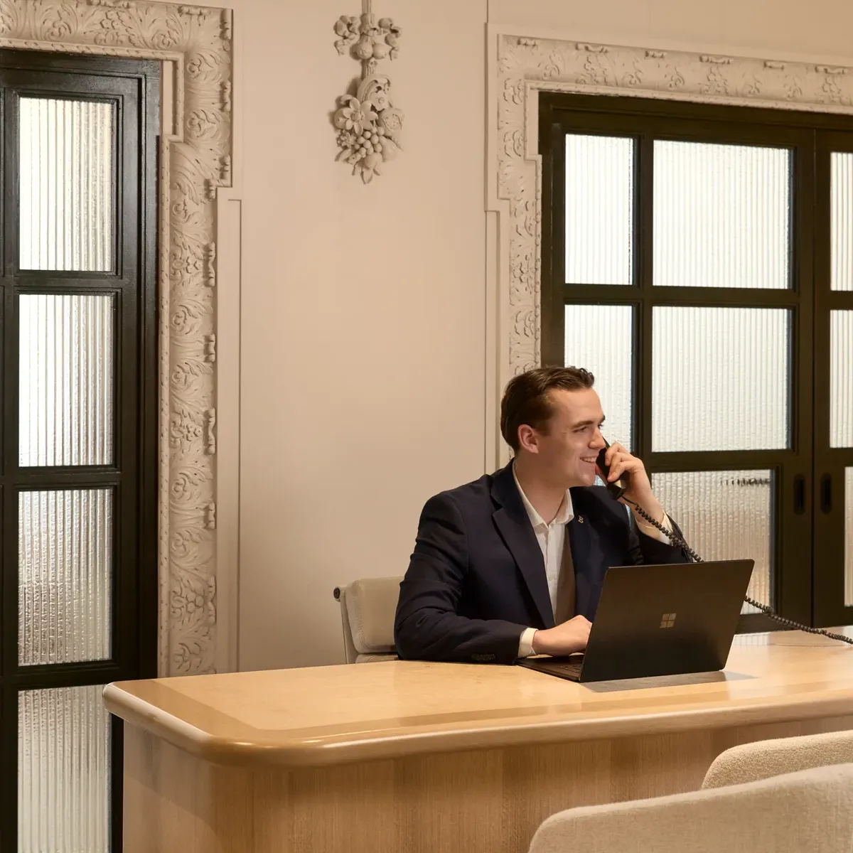 Man sits at a wooden desk using a laptop and phone, in an elegant office with decorative walls and glass doors.