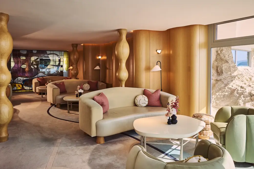 Stylish lounge with curved cream sofas, round tables, and soft pink cushions, set against sculptural wooden columns and a stained-glass artwork.