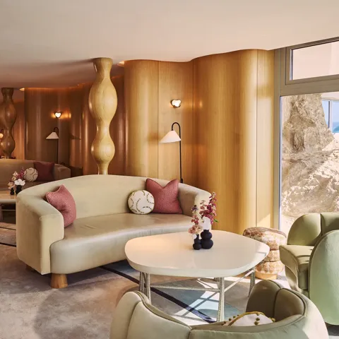 Salon élégant avec canapés crème incurvés, tables rondes et coussins roses, mis en valeur par des colonnes sculpturales en bois et une œuvre en vitrail coloré.