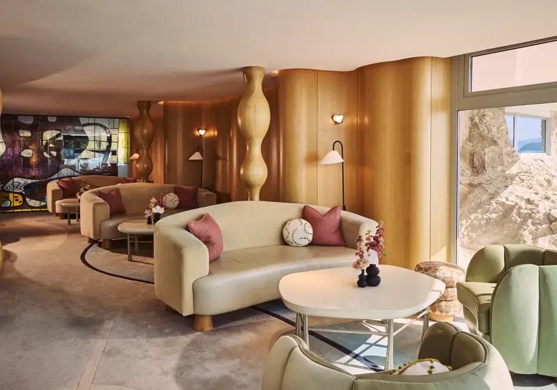 Salon élégant avec canapés crème incurvés, tables rondes et coussins roses, mis en valeur par des colonnes sculpturales en bois et une œuvre en vitrail coloré.