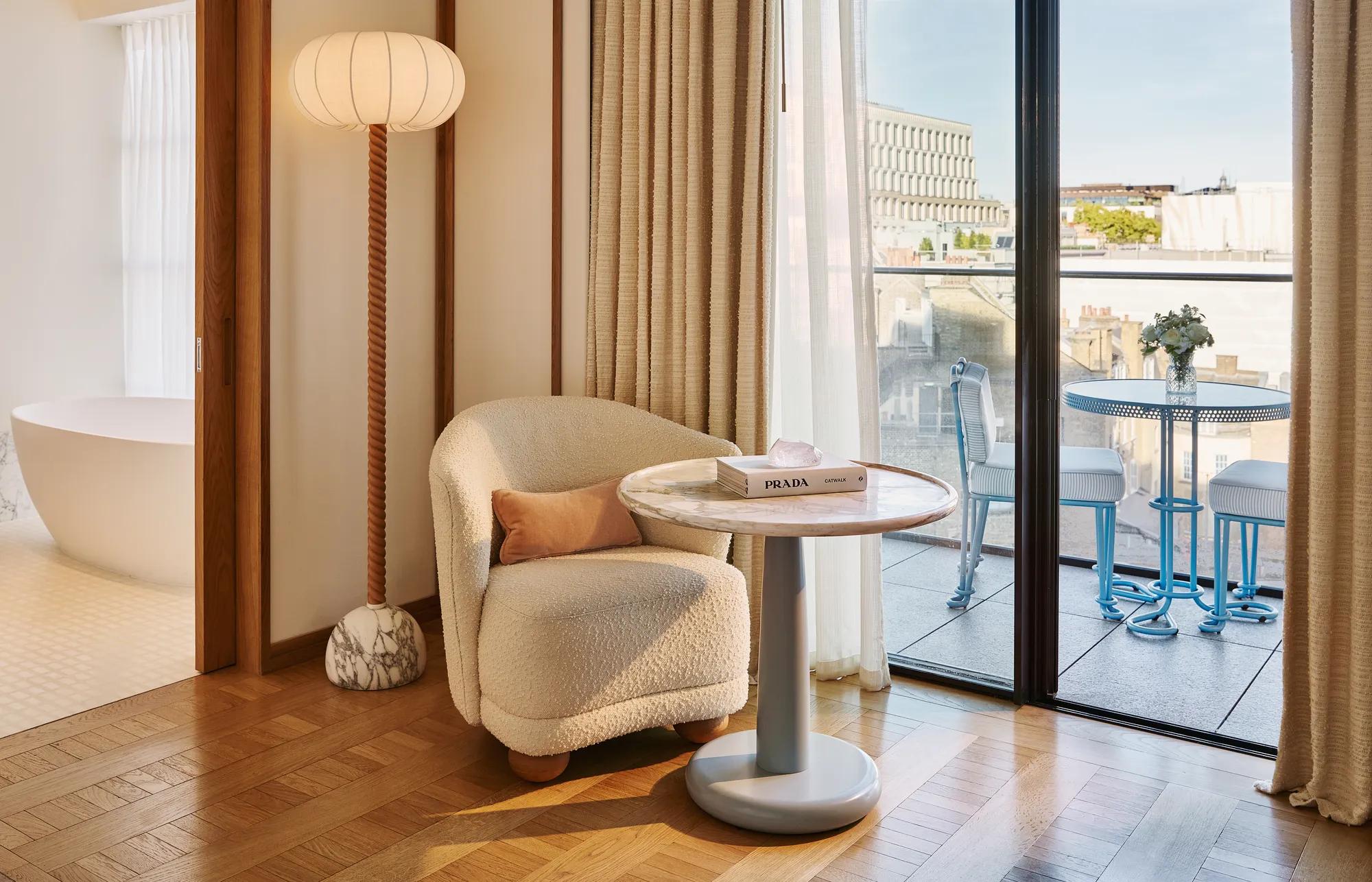Armchair and side table beside floor-to-ceiling doors opening onto a balcony, with freestanding bath visible in the background.