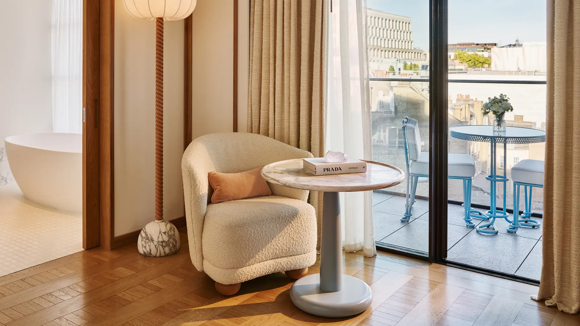 Armchair and side table beside floor-to-ceiling doors opening onto a balcony, with freestanding bath visible in the background.