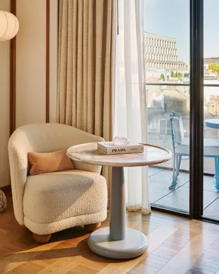 Armchair and side table beside floor-to-ceiling doors opening onto a balcony, with freestanding bath visible in the background.