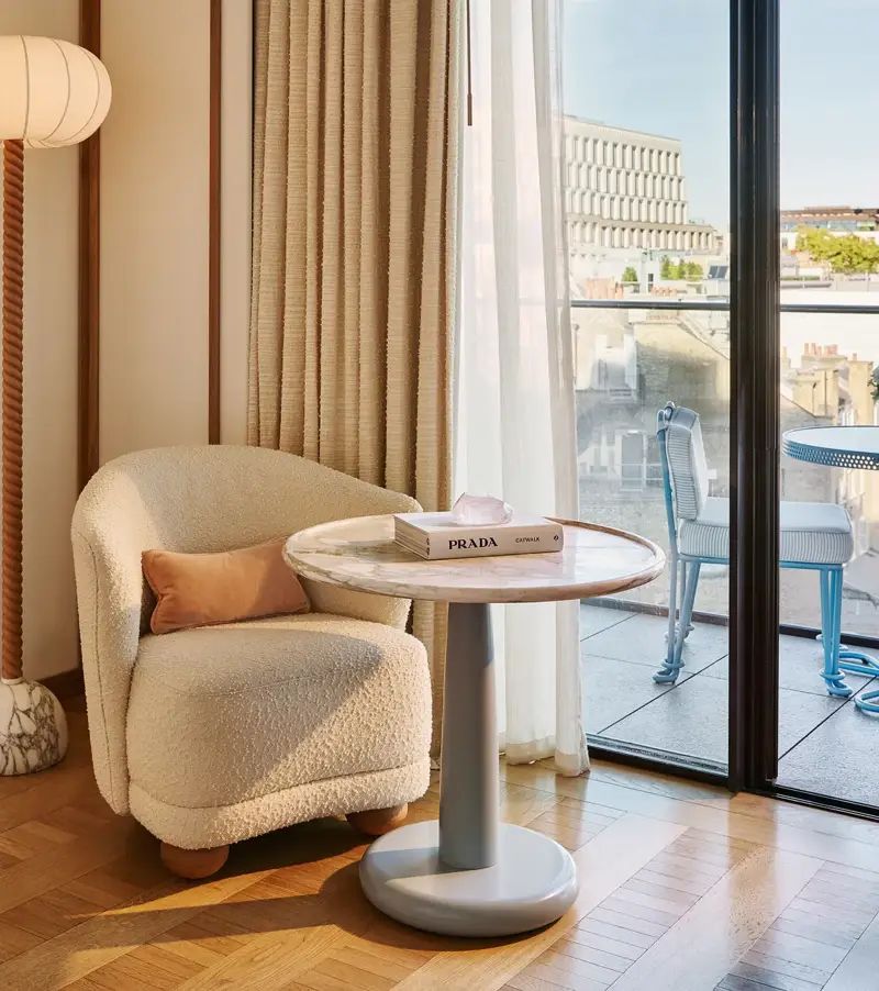 Armchair and side table beside floor-to-ceiling doors opening onto a balcony, with freestanding bath visible in the background.