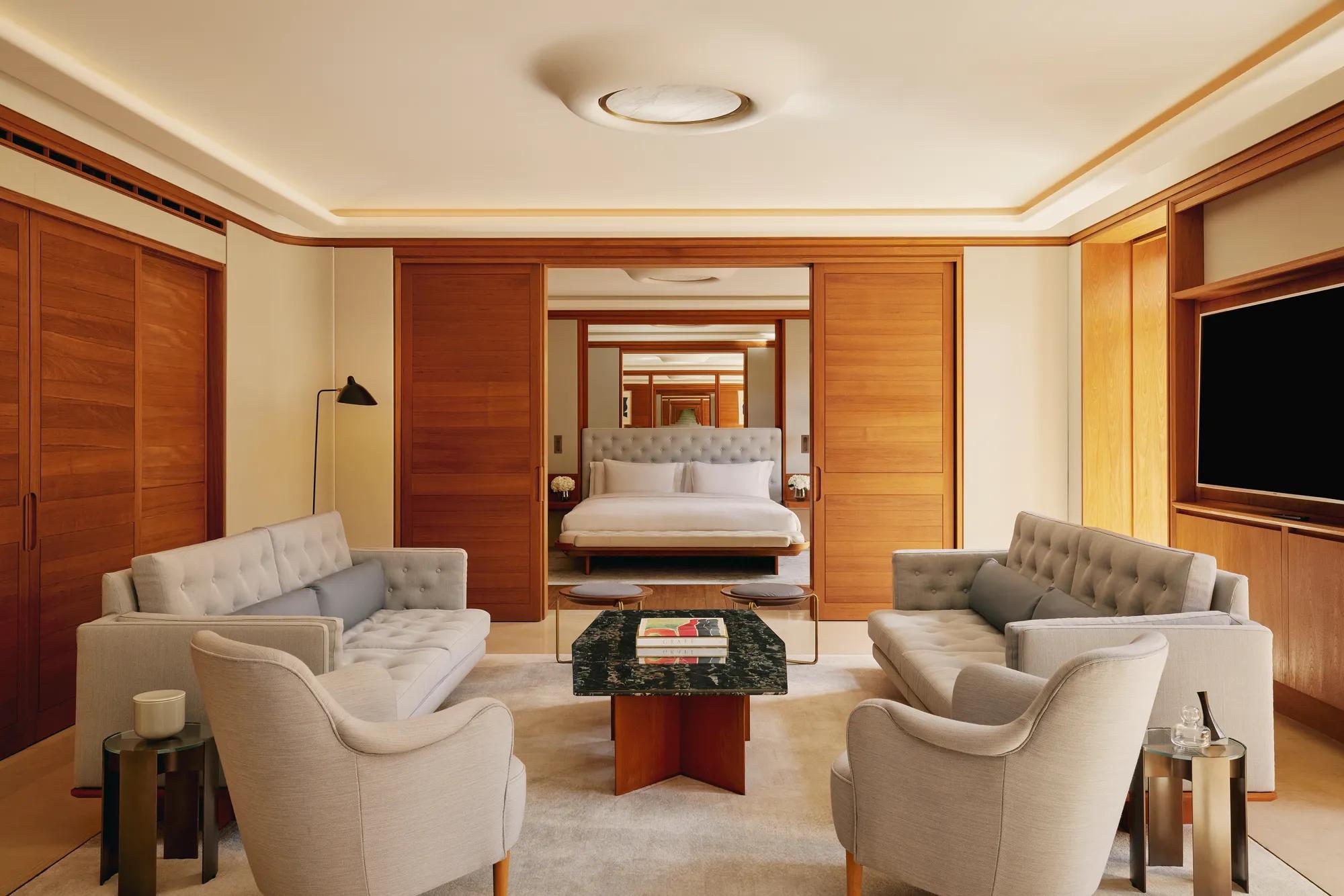 Elegant lounge with sofas and armchairs facing a marble coffee table, opening to a bedroom with a tufted headboard and crisp white bedding.