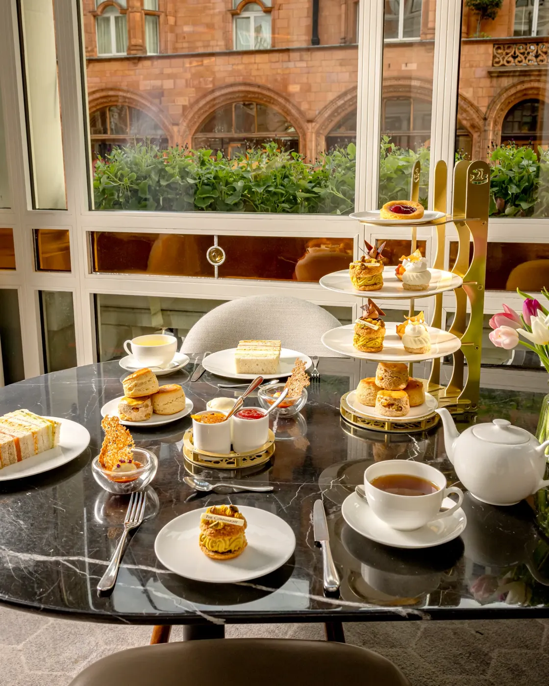 Afternoon tea with tiered pastries, sandwiches, scones, and tea set on a marble table by a window.
