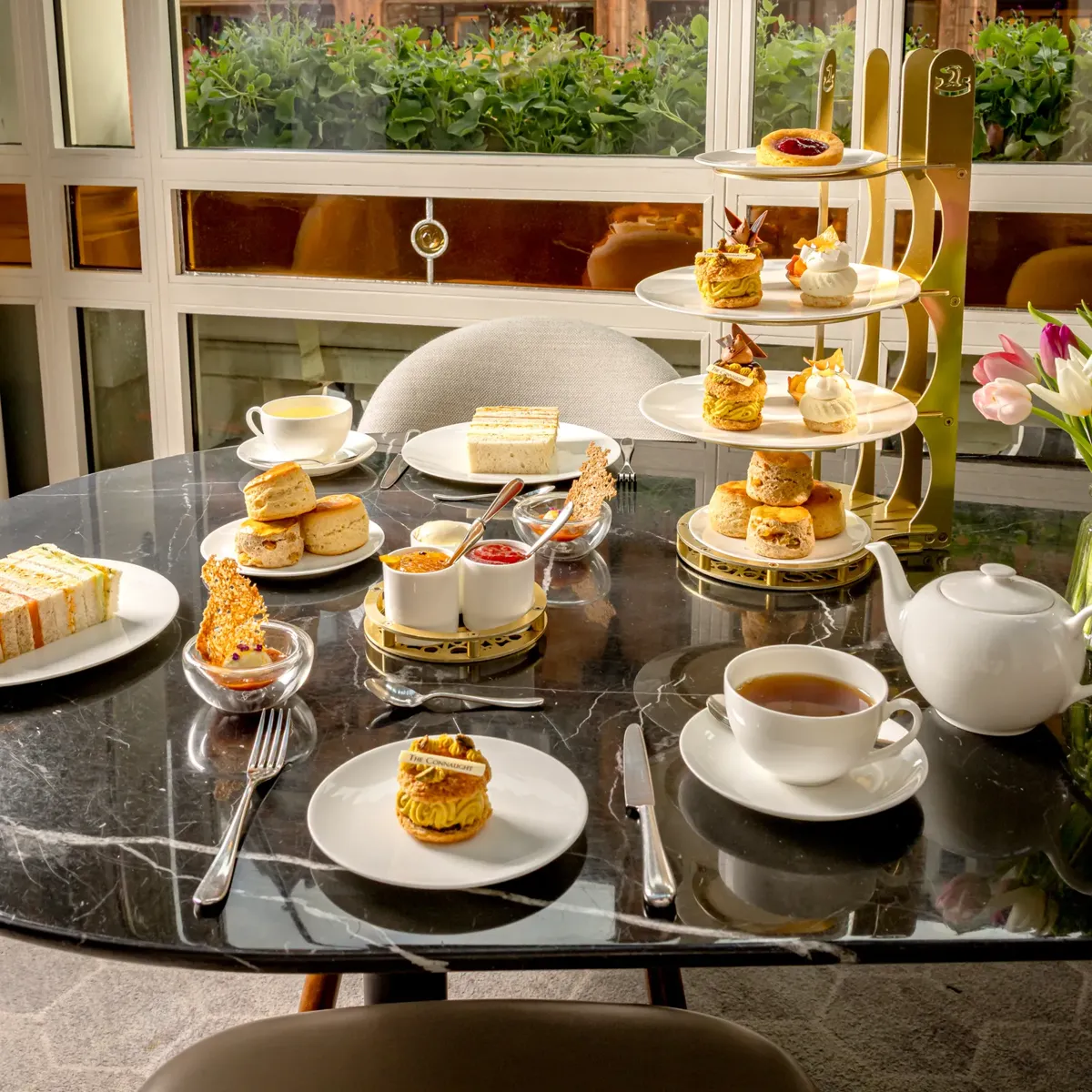 Afternoon tea with tiered pastries, sandwiches, scones, and tea set on a marble table by a window.