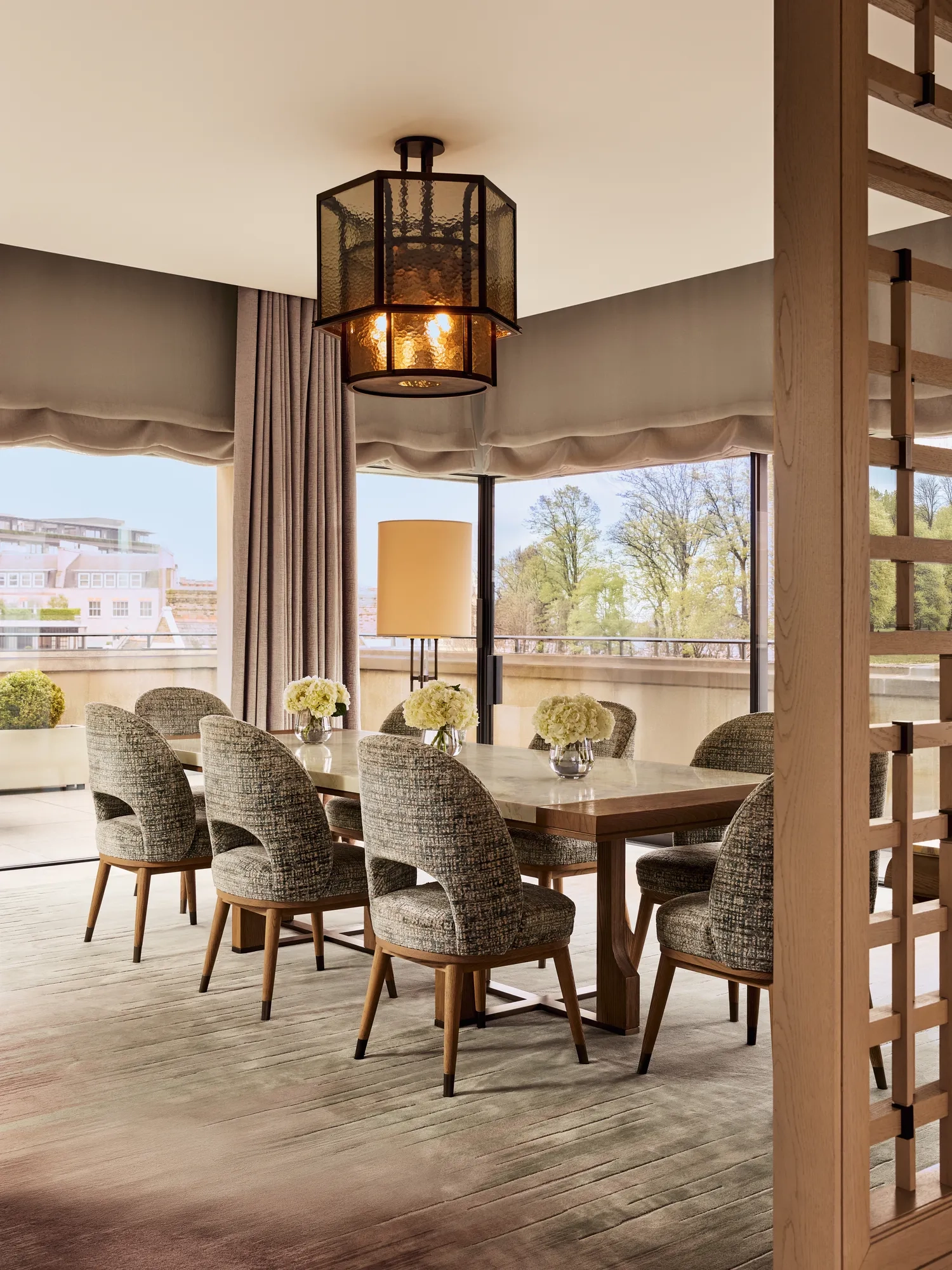 Elegant dining area with six upholstered chairs, wooden table, and large windows overlooking terrace with potted plants.