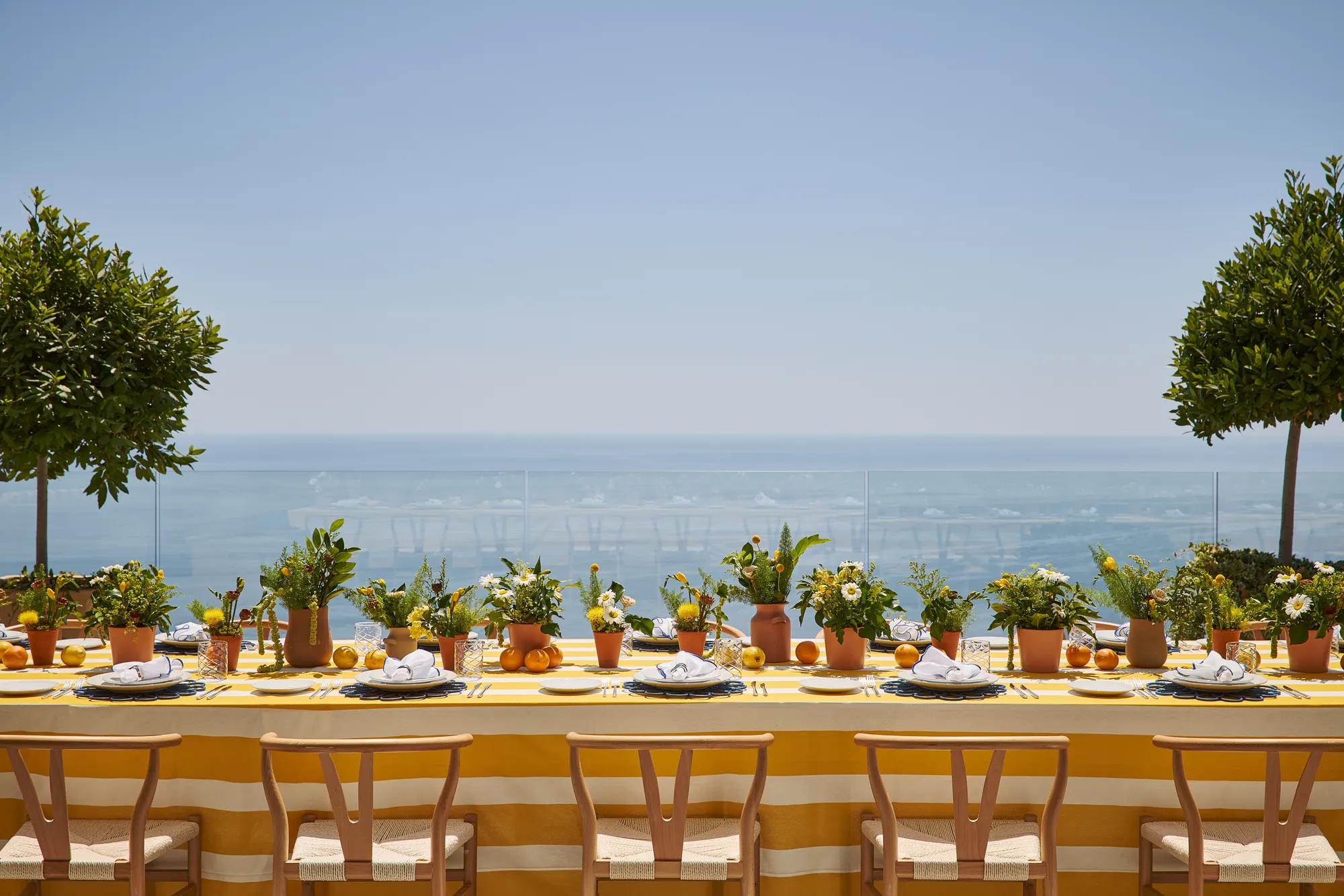 Longue table extérieure avec nappe rayée jaune, pots de plantes et agrumes, surplombant une mer bleue sous un ciel ensoleillé.