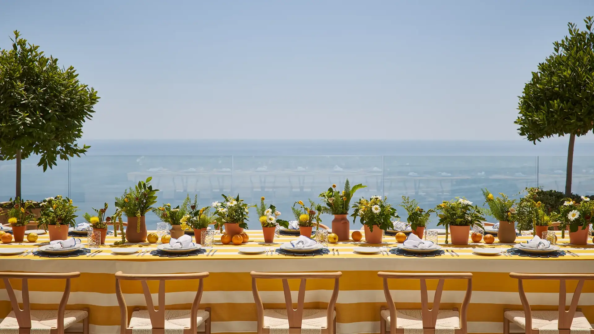 Longue table extérieure avec nappe rayée jaune, pots de plantes et agrumes, surplombant une mer bleue sous un ciel ensoleillé.