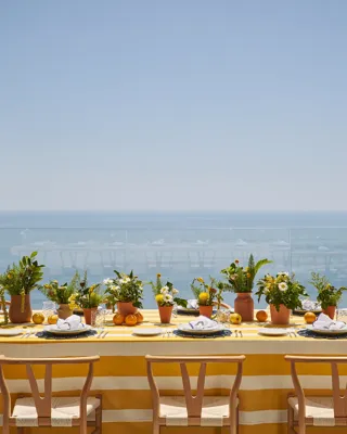 Longue table extérieure avec nappe rayée jaune, pots de plantes et agrumes, surplombant une mer bleue sous un ciel ensoleillé.