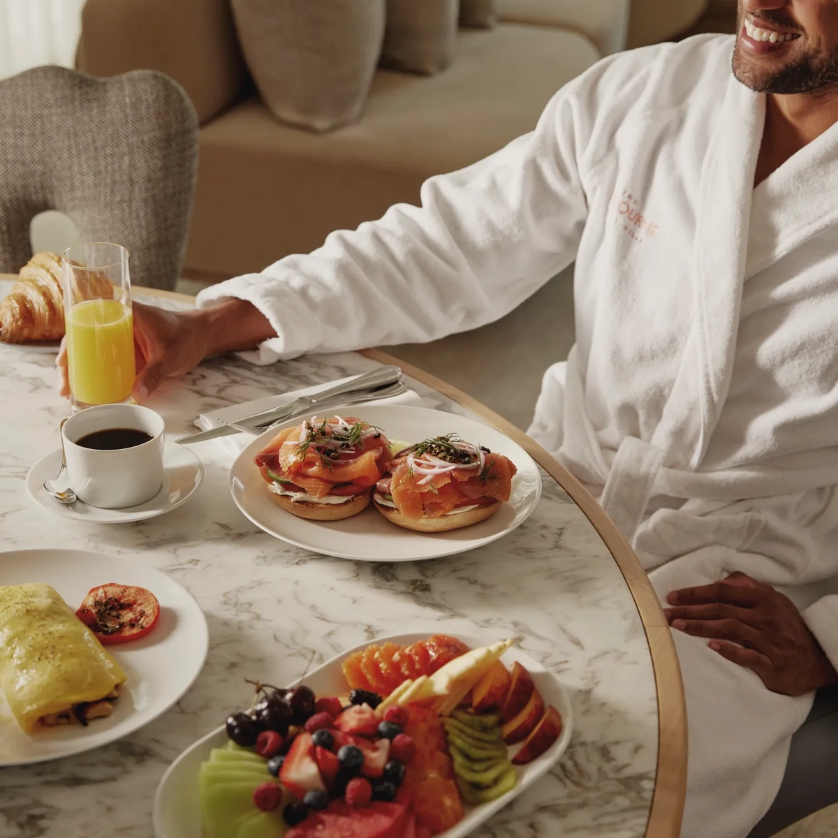 Man in robe enjoying smoked salmon bagels, fresh fruit, and coffee for in-room breakfast at The Maybourne Beverly Hills.