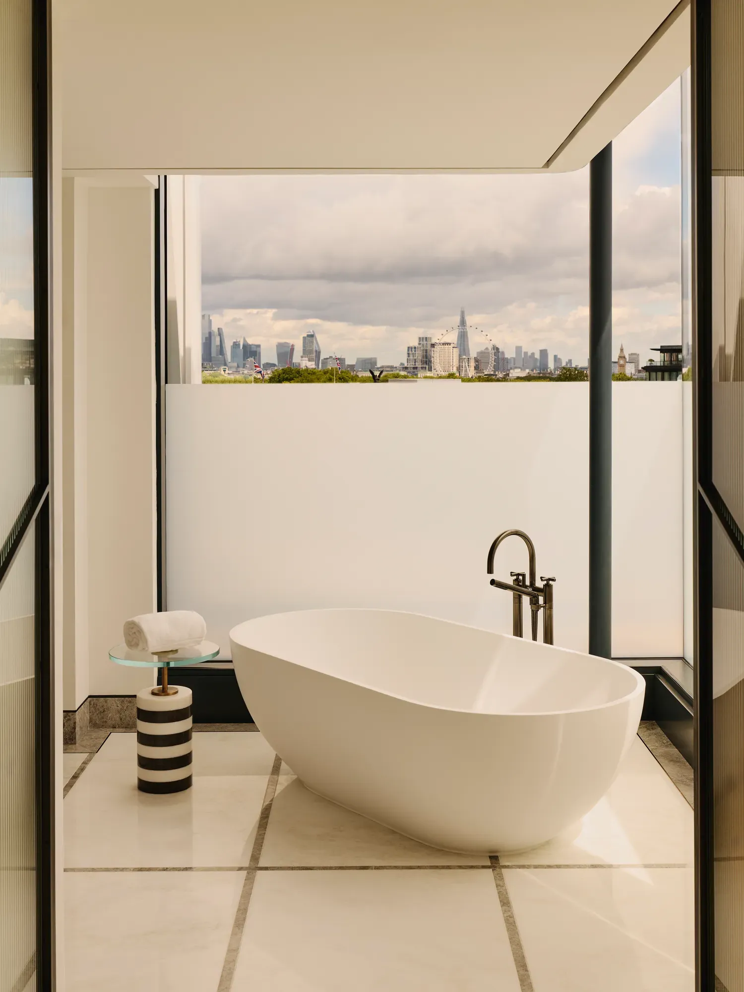 Modern bathroom with freestanding oval bathtub, floor-to-ceiling windows, and city skyline view beyond a private terrace.