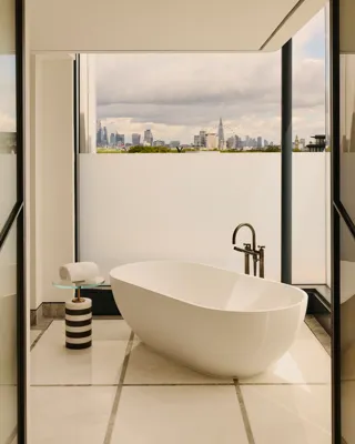 Modern bathroom with freestanding oval bathtub, floor-to-ceiling windows, and city skyline view beyond a private terrace.