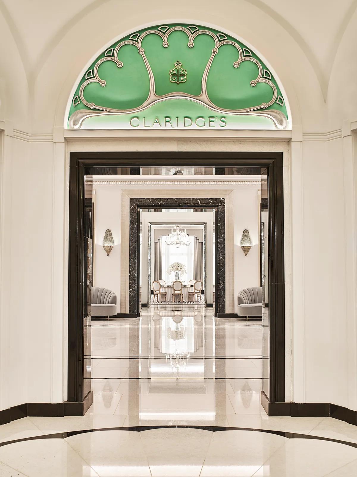 Entrance view of Claridge’s Ballroom corridor, framed by a black and white marble archway and an arched green glass panel with the Claridge’s logo, leading to mirrored walls and chandeliers beyond.