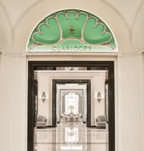Entrance view of Claridge’s Ballroom corridor, framed by a black and white marble archway and an arched green glass panel with the Claridge’s logo, leading to mirrored walls and chandeliers beyond.