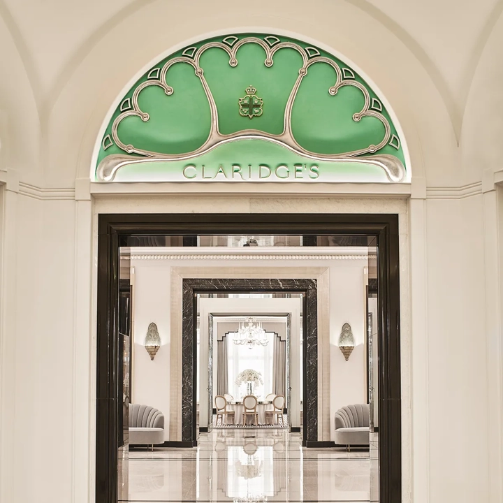 Entrance view of Claridge’s Ballroom corridor, framed by a black and white marble archway and an arched green glass panel with the Claridge’s logo, leading to mirrored walls and chandeliers beyond.