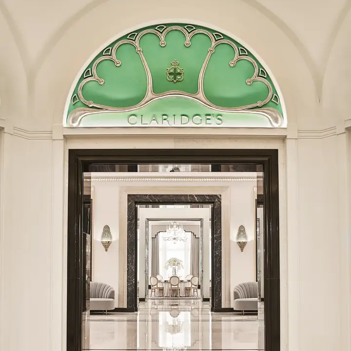 Entrance view of Claridge’s Ballroom corridor, framed by a black and white marble archway and an arched green glass panel with the Claridge’s logo, leading to mirrored walls and chandeliers beyond.