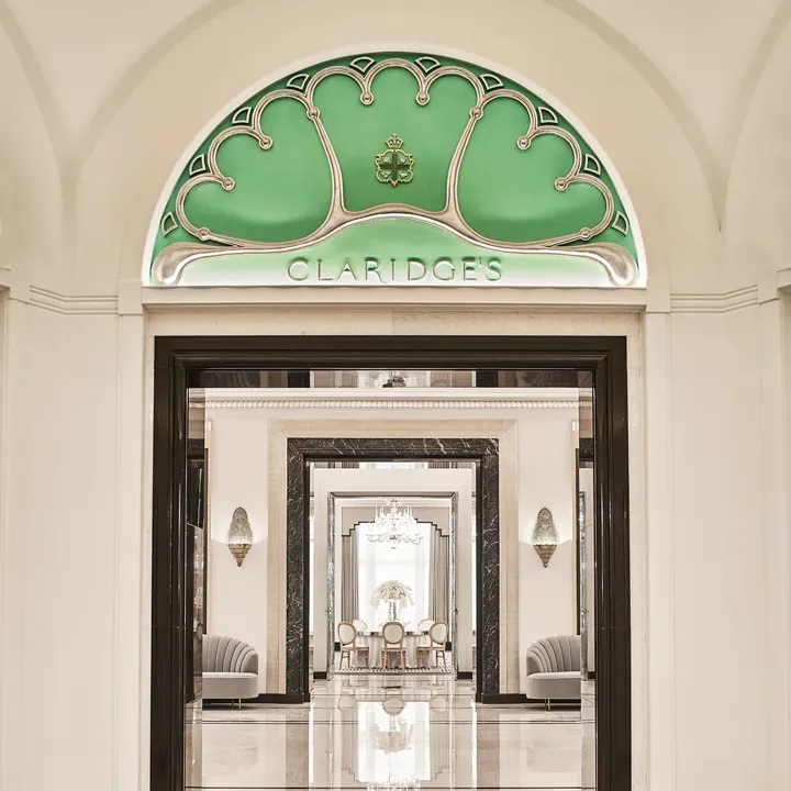 Entrance view of Claridge’s Ballroom corridor, framed by a black and white marble archway and an arched green glass panel with the Claridge’s logo, leading to mirrored walls and chandeliers beyond.
