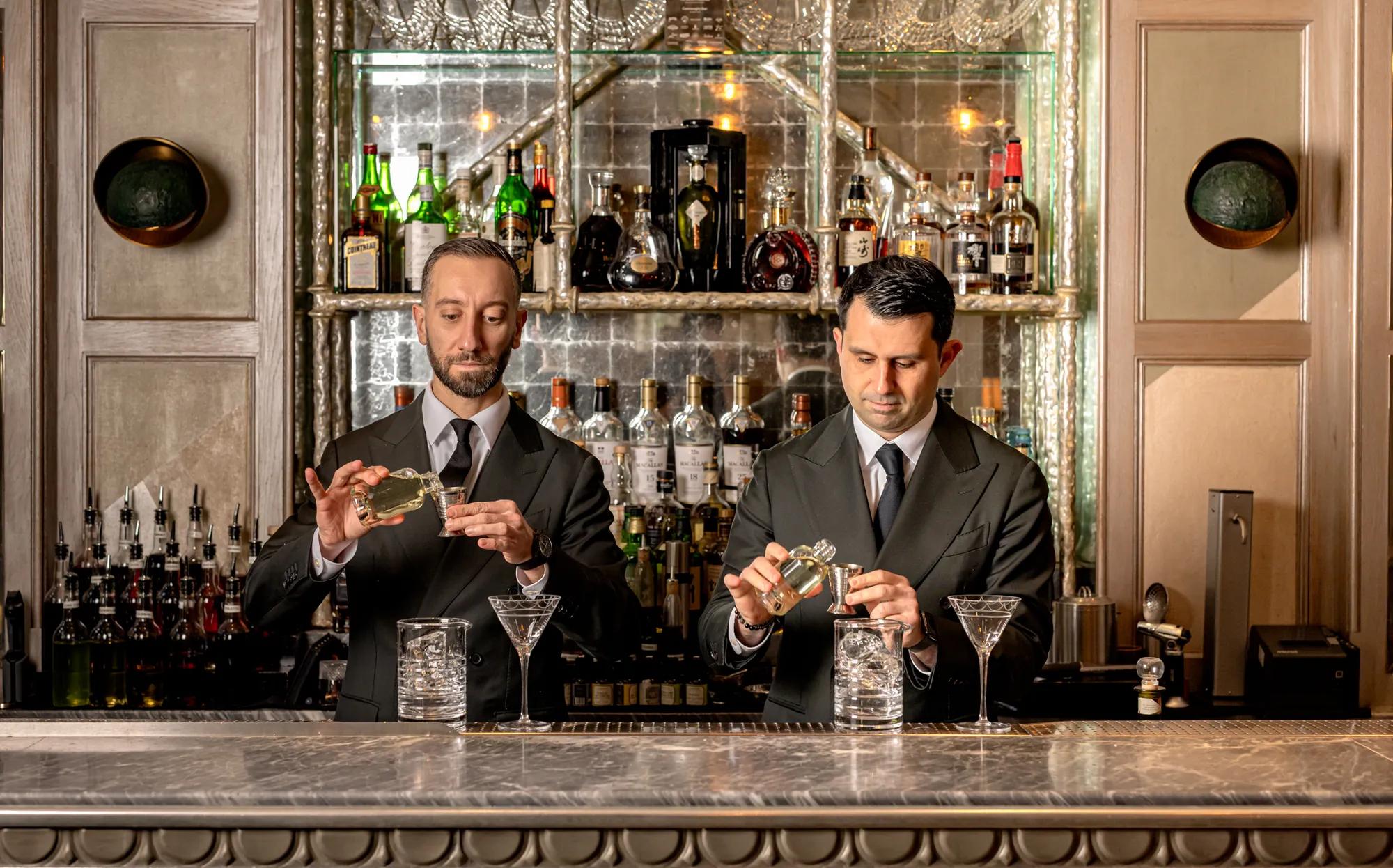 **Alt text (WCAG 2.2 AA, under 125 characters):**

Two bartenders prepare cocktails behind a marble bar, with shelves of spirits and glassware in the background.
