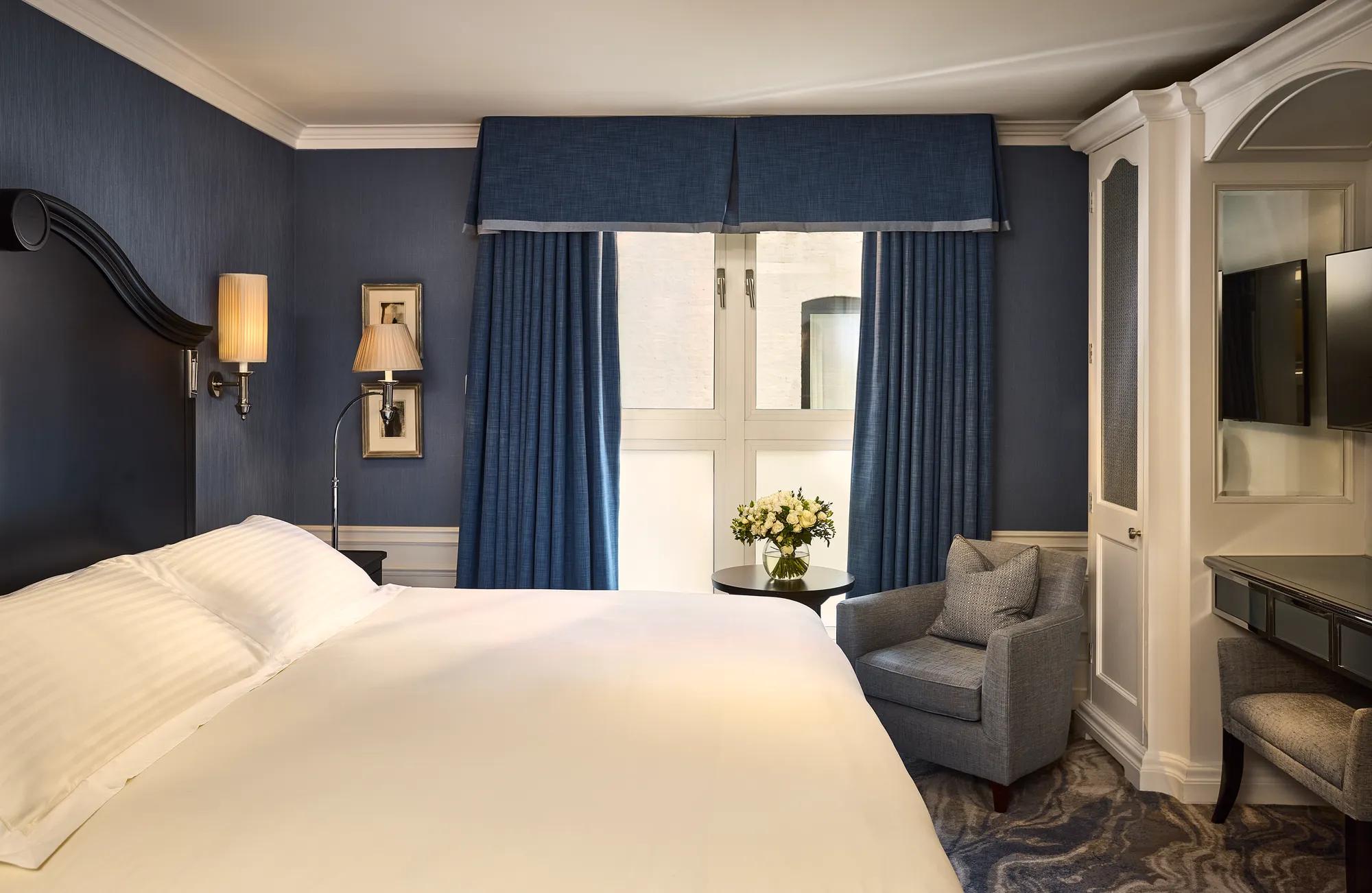 Elegant guest room with king bed, blue walls, armchair by window, and dressing table.