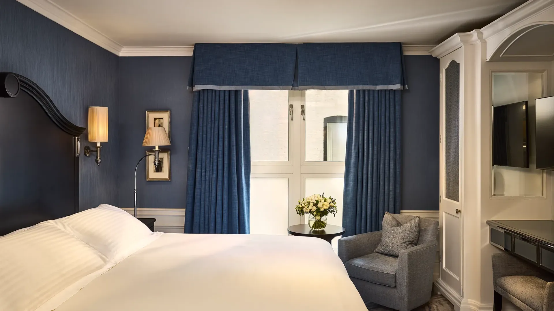 Elegant guest room with king bed, blue walls, armchair by window, and dressing table.
