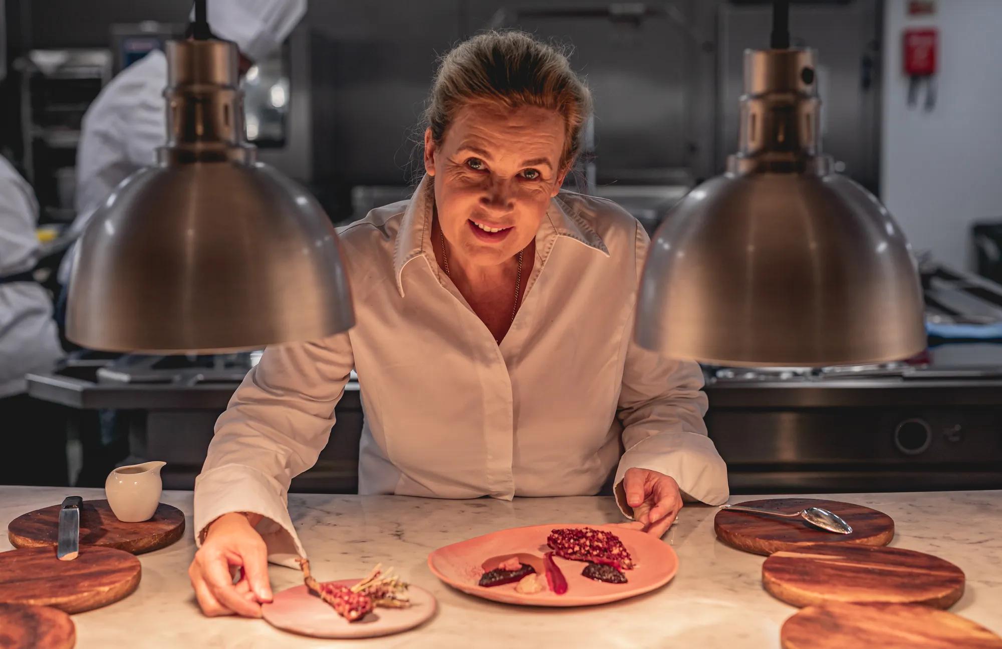 Chef plating dessert at marble counter under heat lamps in professional kitchen.