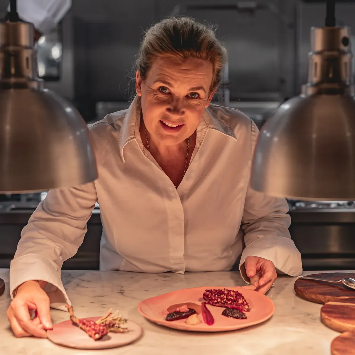 Chef plating dessert at marble counter under heat lamps in professional kitchen.