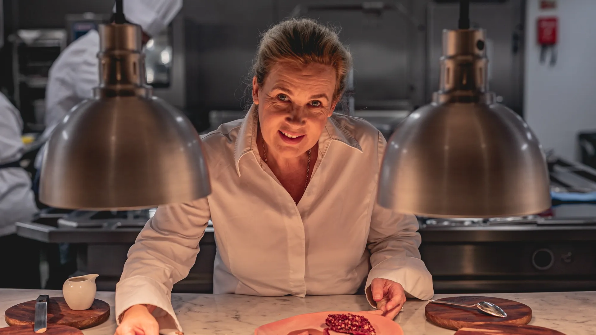 Chef plating dessert at marble counter under heat lamps in professional kitchen.