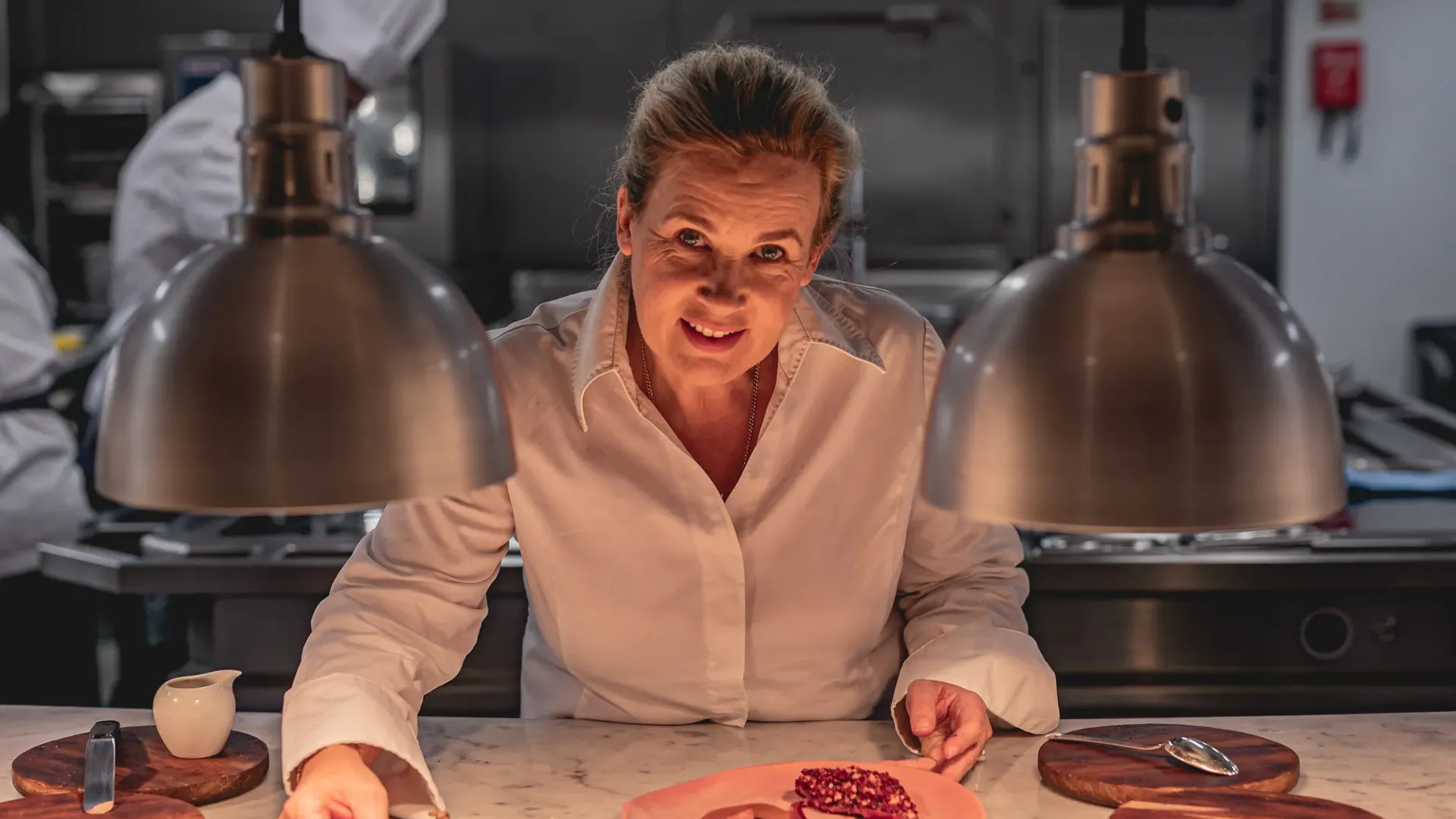 Chef plating dessert at marble counter under heat lamps in professional kitchen.