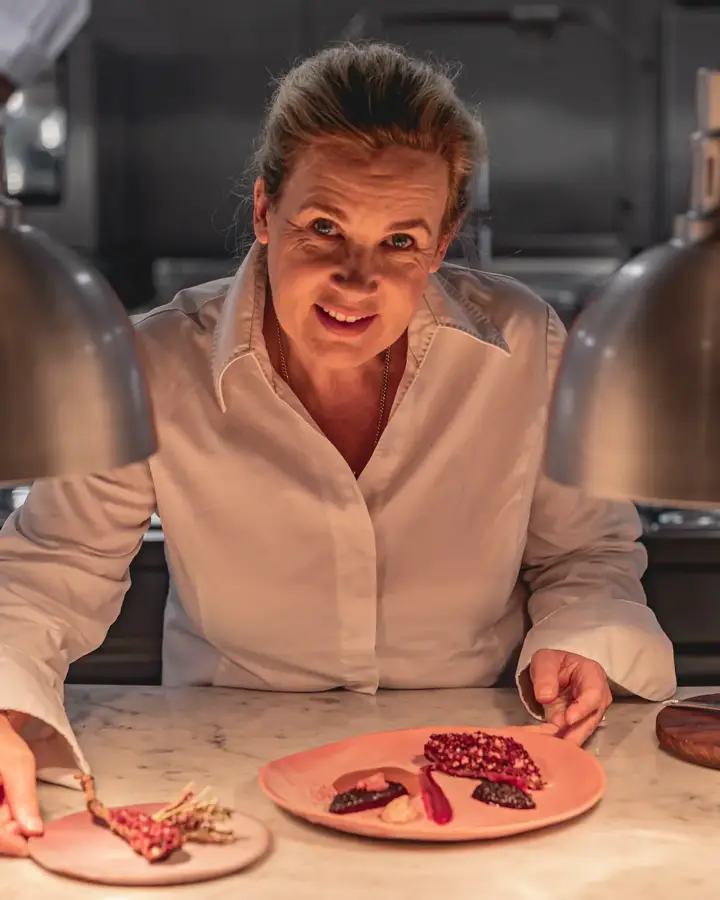 Chef plating dessert at marble counter under heat lamps in professional kitchen.