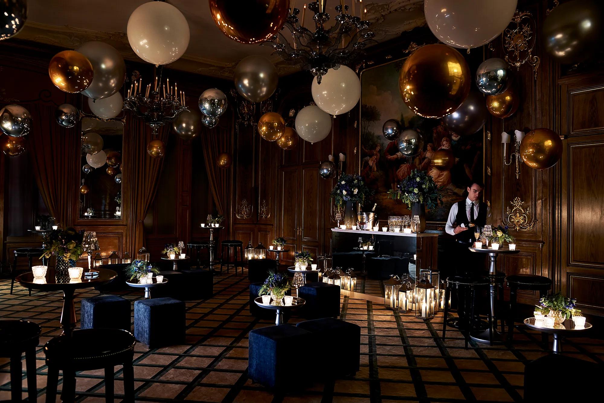 Dimly lit Claridge’s ballroom decorated for an evening celebration with gold, silver, and white balloons, flickering candles, velvet seating, and a bartender preparing drinks beneath a large classical painting.