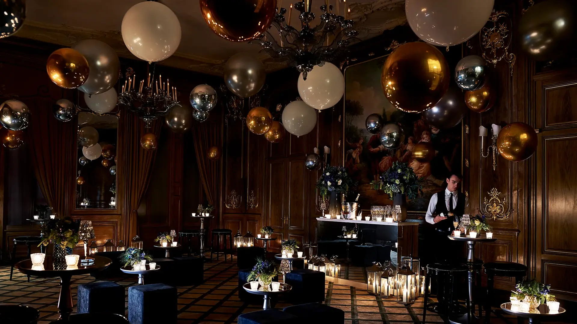Dimly lit Claridge’s ballroom decorated for an evening celebration with gold, silver, and white balloons, flickering candles, velvet seating, and a bartender preparing drinks beneath a large classical painting.