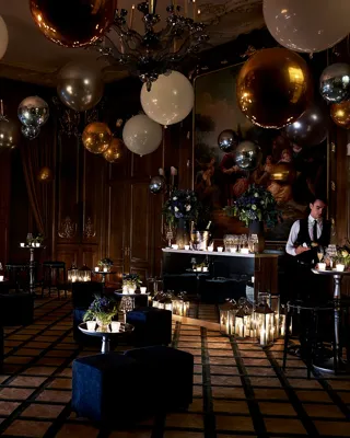 Dimly lit Claridge’s ballroom decorated for an evening celebration with gold, silver, and white balloons, flickering candles, velvet seating, and a bartender preparing drinks beneath a large classical painting.