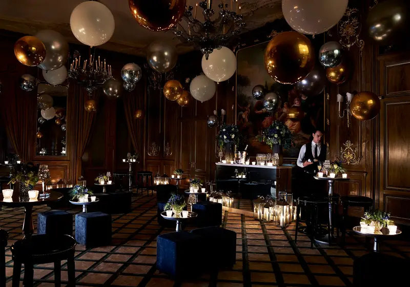 Dimly lit Claridge’s ballroom decorated for an evening celebration with gold, silver, and white balloons, flickering candles, velvet seating, and a bartender preparing drinks beneath a large classical painting.