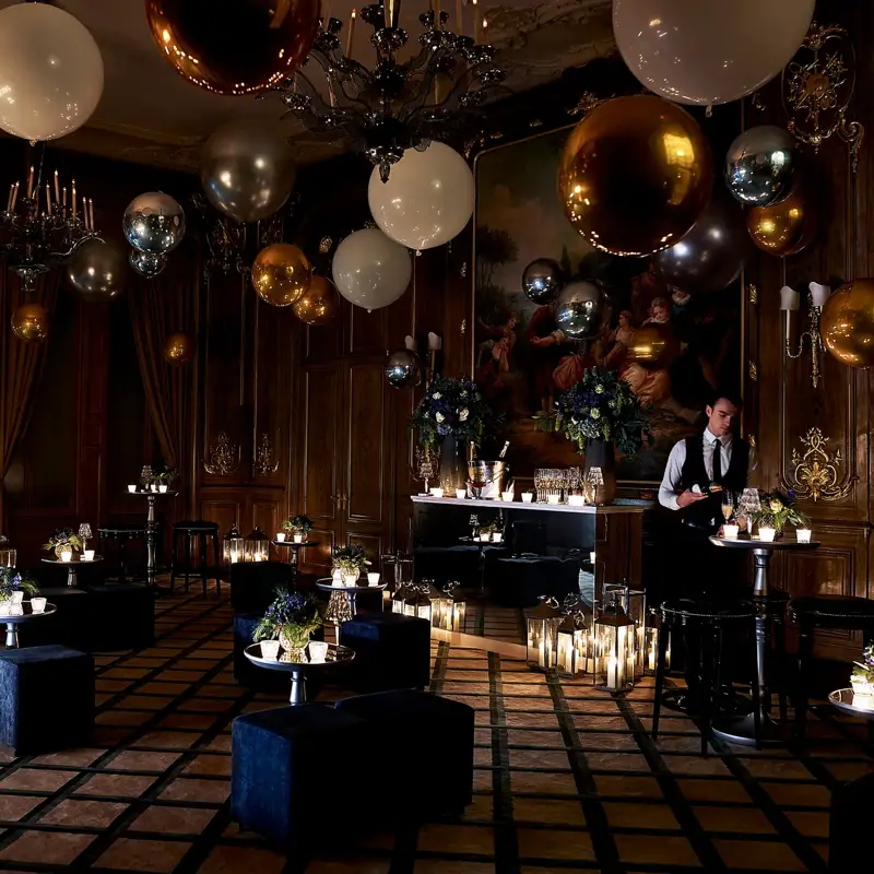 Dimly lit Claridge’s ballroom decorated for an evening celebration with gold, silver, and white balloons, flickering candles, velvet seating, and a bartender preparing drinks beneath a large classical painting.