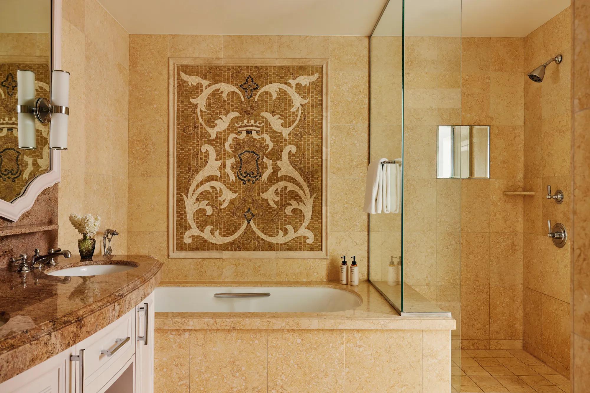 Elegant bath showing an ornate mosaic panel above the tub, stone vanity, and a glass‑enclosed walk‑in shower with chrome fixtures; limestone tile throughout.