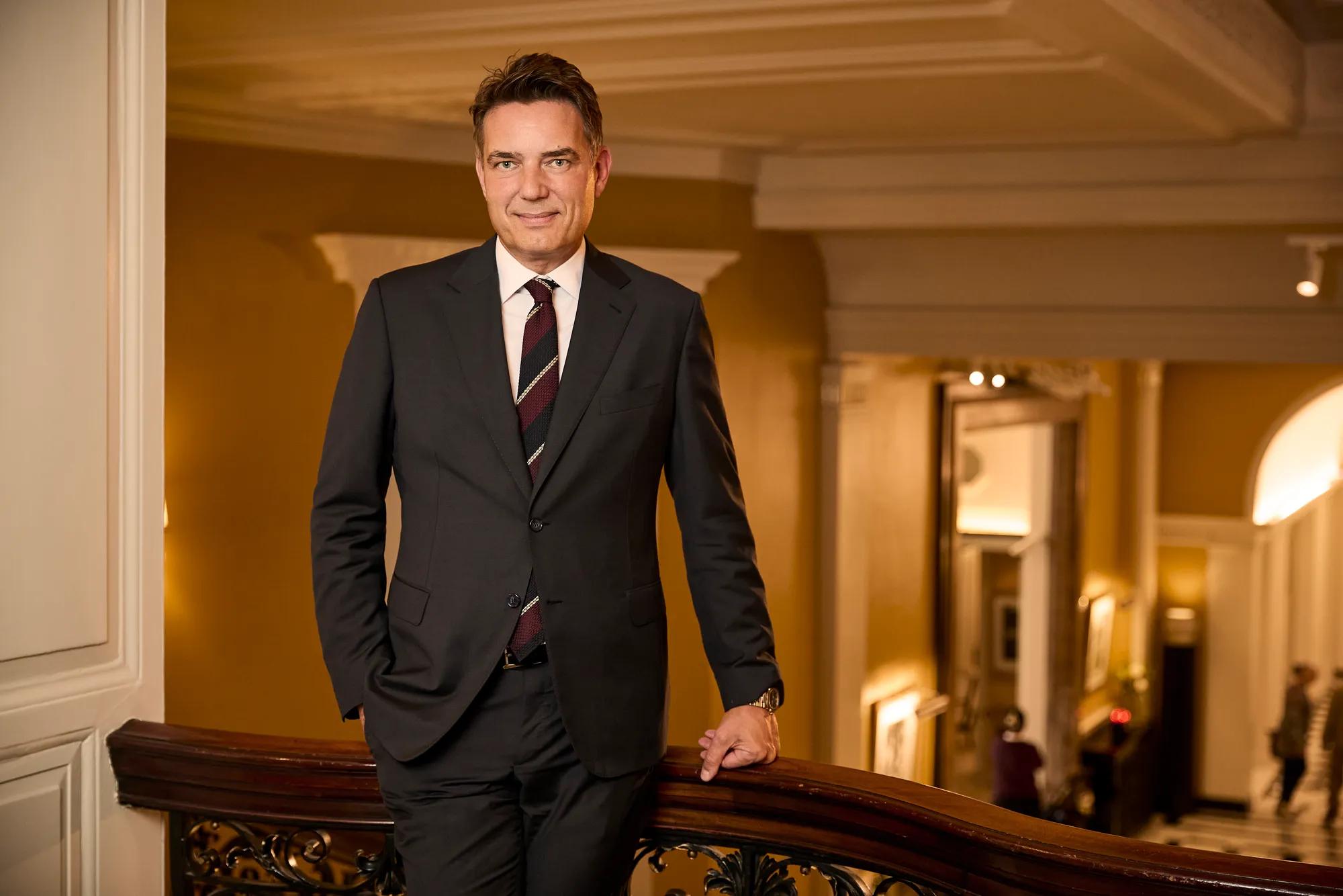 Man in a dark suit leaning on a staircase railing in an elegant, warmly lit hotel interior