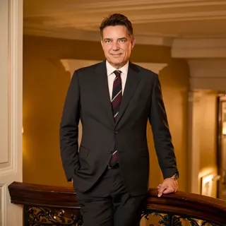 Man in a dark suit leaning on a staircase railing in an elegant, warmly lit hotel interior