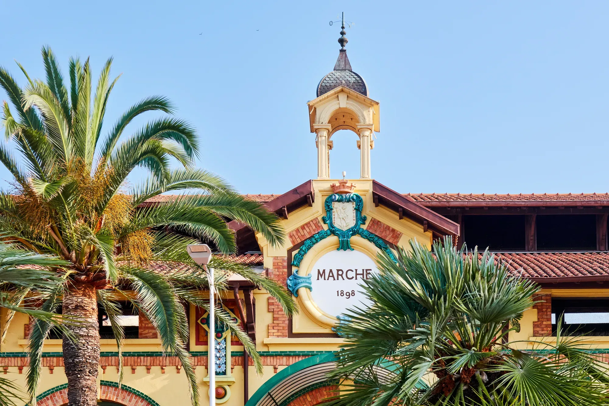 Façade historique de marché avec clocher et palmiers, ornée d’un blason décoratif sous un ciel bleu.