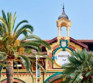 Historic market façade with clock tower and palm trees, featuring a decorative crest under a clear blue sky.