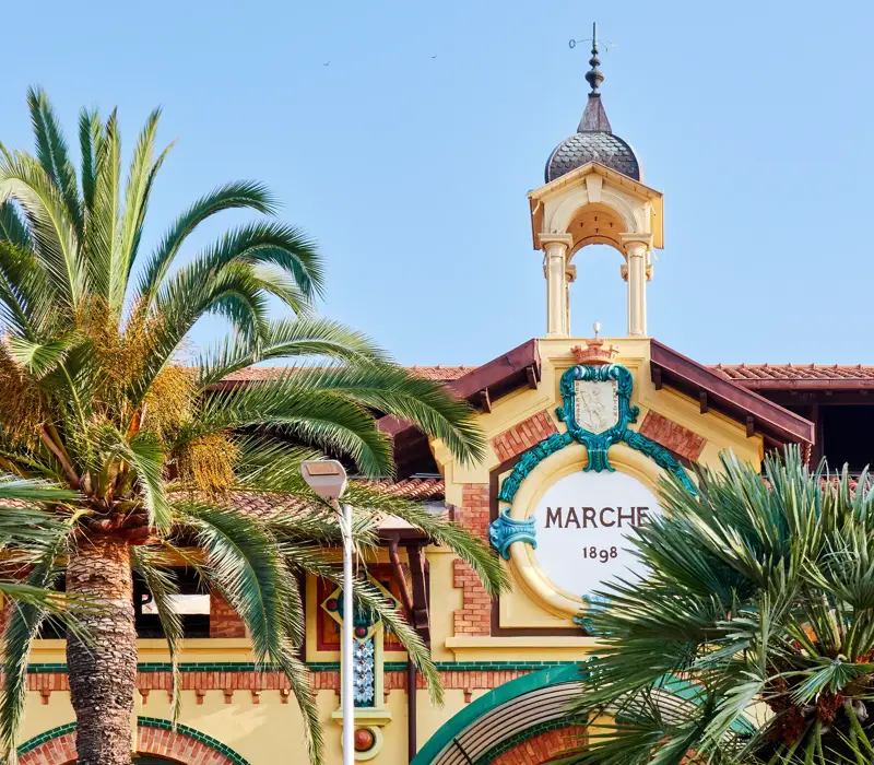 Historic market façade with clock tower and palm trees, featuring a decorative crest under a clear blue sky.