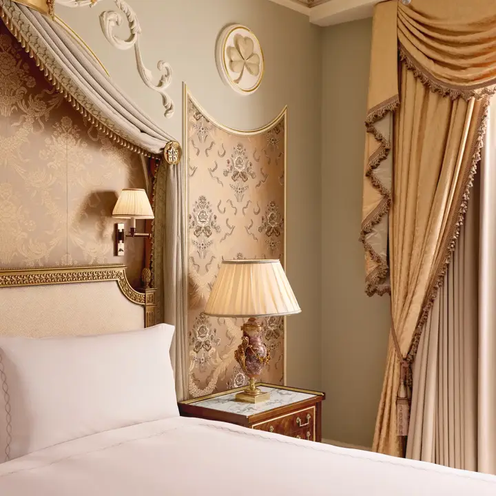 Ornate bedroom with canopy bed, gold-patterned headboard, bedside lamp on wooden table, and draped curtains beneath decorative mouldings.